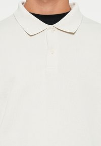 Tricou polo alb confecționat din țesătură striată, având un guler clasic și un placket cu trei nasturi. Suprafață texturată cu o formă ajustată.