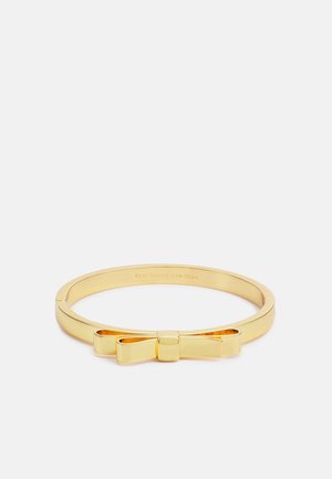 Bracelet en métal doré avec un détail en forme de nœud sur le dessus, élégant et poli, reposant sur un fond blanc uni.