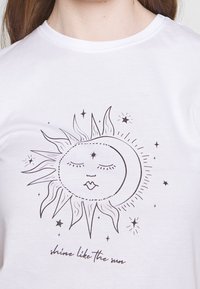 Camiseta blanca con un diseño de arte lineal negro de un sol y una luna con estrellas, acompañado del texto "brilla como el sol" debajo.