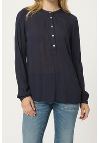 Femme portant un chemisier bleu marine à manches longues avec cinq boutons et un jean bleu clair, debout les mains détendues le long du corps.