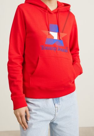 Henkilö, joka käyttää punaista hupparia, jossa on sinivalkoinen tähtilogo ja teksti "Tommy Jeans", yhdistettynä vaaleansinisiin farkkuihin.