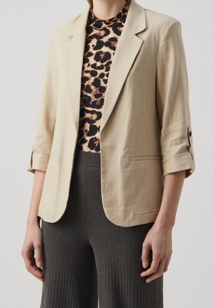 Kvinde iført beige blazer med opkørte ærmer, leopardprint top og mørkegrå ribstrikkede vidde ben bukser, stående mod en neutral baggrund.