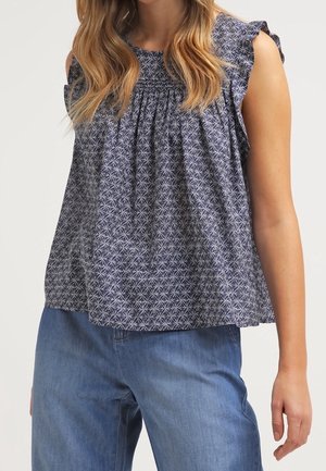 Vrouw draagt een mouwloze marineblauwe blouse met patroon, gerimpelde schouders en losse blauwe spijkerbroek, staande tegen een effen achtergrond.