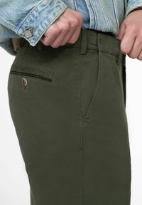 Pantaloni verde oliva a vestibilità slim, realizzati in un tessuto liscia. Presentano una tasca anteriore, una tasca posteriore con bottone e una chiusura con zip.