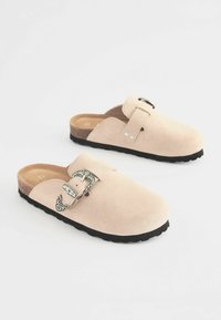 Sabots en daim rose clair avec semelle en caoutchouc noir, strap ajustable avec boucle en métal, et texture lisse. Pas de motifs ou d'accents visibles.
