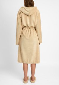 Beżowy szlafrok z kapturem wykonany z teksturowanej tkaniny, z paskiem w talii i długimi rękawami. Miękkie, puszyste kapcie dopełniają wygląd.