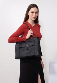 Black leather tote bag with a front flap, gold hardware, and dual shoulder straps. Smooth texture with a structured design.
