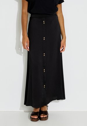 Femme debout portant une longue jupe noire avec une rangée de petits boutons beiges sur le devant et des sandales compensées noires.