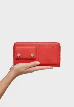 Mano che tiene un portafoglio rosso con chiusura lampo completa, tasca con bottone a scatto per monete e scomparto per carte, impresso con il nome del marchio TOUS.
