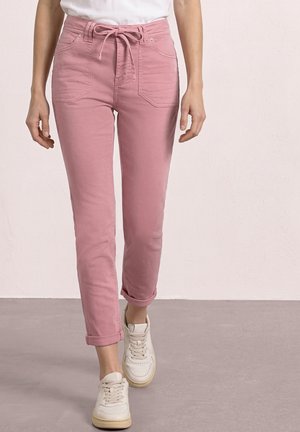 Osoba nosząca spodnie z wysokim stanem w kolorze przybrudzonego różu z przednimi kieszeniami i paskiem do wiązania, w komplecie z podwiniętymi mankietami i białymi trampkami, stojąca na szarej podłodze.