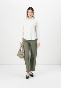 Woman standing wearing a white button-up shirt, olive green pants, black pointed shoes, and holding a snakeskin patterned handbag.