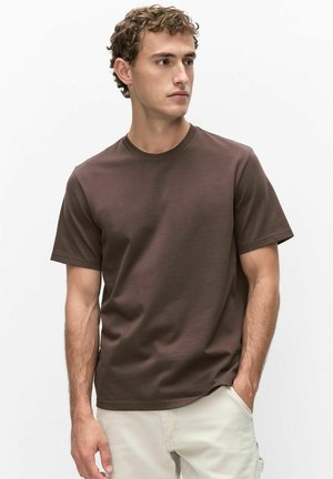Ung mand med krøllet hår iført en brun t-shirt med korte ærmer og lyse beige bukser, som kigger til siden med den ene hånd i lommen.