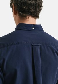 Marineblauwe shirt met knoopsluiting op de kraag, voorzien van een split aan de achterkant en een klein wit knoopdetail op de kraag. Gladde textuur.