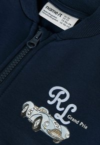 Tumši zili rāvējslēdzēja apģērbs ar vintage sacīkšu automašīnas grafiku "RL Grand Prix" un apģērba etiķeti, kurā norādīts lielums un teksts "Ražots Bangladešā".