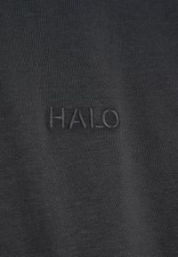 Svart bomullstyg med en broderad "HALO"-logotyp i en matchande mörk tråd som skapar ett subtilt och texturerat utseende.