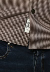 Bruine geklede shirt met een gestructureerde afwerking en een zwarte knoop, met een label dat "DEELUXE" leest. Draagt comfortabel met een donkere spijkerbroek.