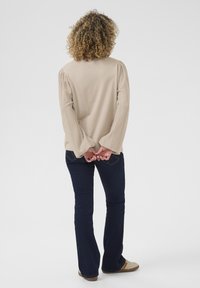 Beige strikket sweater med pufærmer, parret med mørkeblå flared jeans. Personen står med hænderne foldet bag ryggen.