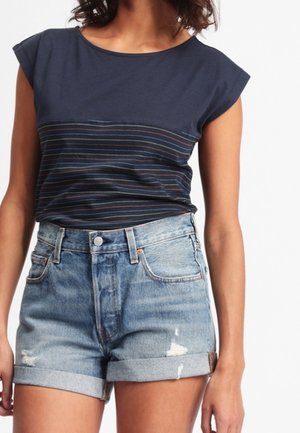 Kobieta nosząca granatowy top bez rękawów z cienkimi kolorowymi paskami oraz jasnoniebieskie podwinięte krótkie spodenki jeansowe z postrzępieniami.