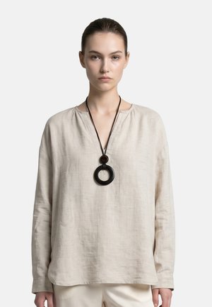 Vrouw draagt een losse lichtbeige top met lange mouwen en een grote zwarte ronde hanger aan een zwarte koord.