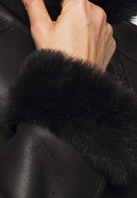 Svart läderjacka med en mjuk, texturerad svart pälskrage och dragkedjedetaljer. Insidan visar en slät yta och skräddarsydd design.