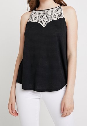 Vrouw draagt een mouwloze zwarte top met een witte kanten geometrische halslijn, gecombineerd met een witte broek tegen een effen achtergrond.