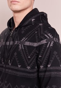 Svart zip-up hoodie med geometriska grå mönster, som har en huva, framdragkedja och textilstruktur. Accentueras av subtila ränder.