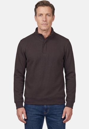 Man draagt een donkerbruine trui met een kwart ritssluiting en blauwe jeans, staand tegen een effen lichte achtergrond.