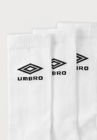 Białe sportowe skarpetki z żebrowaną fakturą, z czarnym logo Umbro na środku i dwoma diamentowymi kształtami nad nazwą marki.