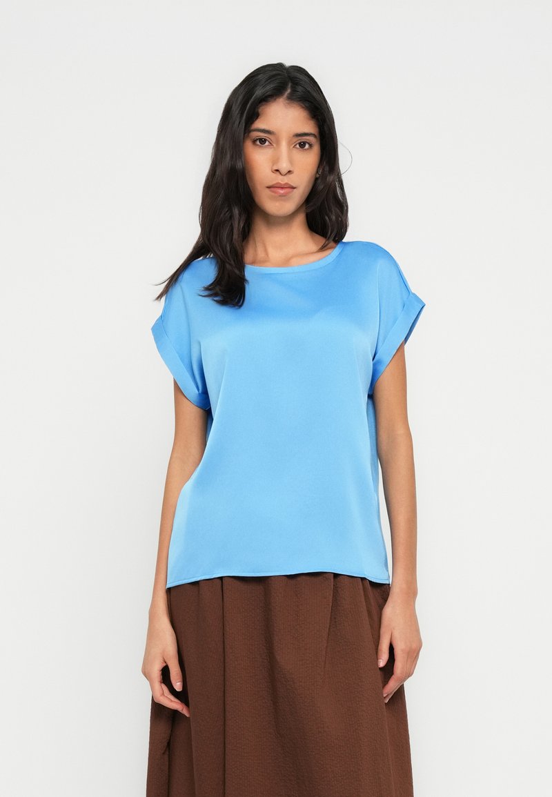 Niebieska, lekka bluzka o luźnym kroju, z podwiniętymi rękawami, gładką teksturą i okrągłym dekoltem, zestawiona z brązową spódnicą o fakturze.