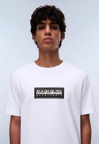 Hvid t-shirt med korte ærmer, der har et sort rektangulært logo med hvid tekst "NAPAPUR" og "geographic" i midten. Glat bomuldsstof.