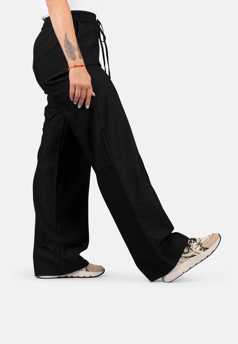 Zwarte, losse broek met een elastische tailleband en trekkoord. Heeft platte naden en rechte pijpen. Draagt met beige en gemêleerde sneakers.