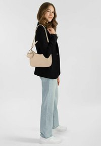 Borsa a tracolla in pelle beige con forma curva, dotata di manico superiore e tracolla regolabile, abbinata a un blazer nero e jeans in denim chiaro.