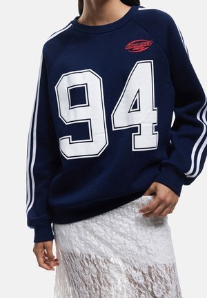 Personne portant un sweat-shirt bleu marine avec des rayures blanches sur les manches, un grand "94" blanc sur le devant, un logo rouge "Unlimited Energy" et une jupe en dentelle blanche.
