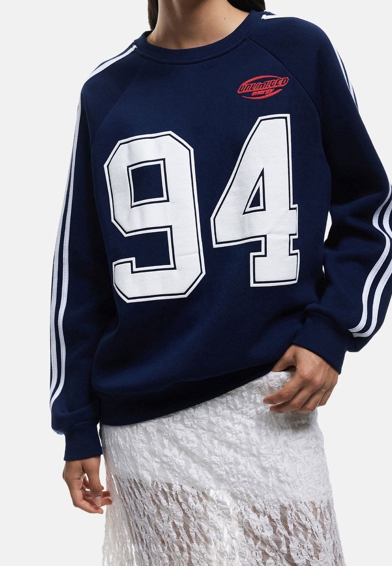 Personne portant un sweat-shirt bleu marine avec des rayures blanches sur les manches, un grand "94" blanc sur le devant, un logo rouge "Unlimited Energy" et une jupe en dentelle blanche.