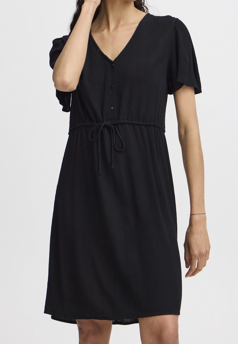 Femme portant une robe noire mi-longue à manches courtes, col en V, boutonnée à l'avant, avec ceinture nouée, sur un fond uni.