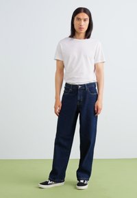 Tumma sininen farkkumateriaali, rento istuvuus, nappisuljenta edessä, yhdistettynä yksinkertaiseen valkoiseen crew-neck t-paitaan ja mustiin lenkkareihin.