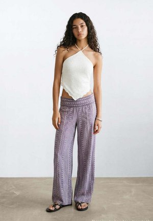 Jonge vrouw met krullend zwart haar die een witte haltertop, paarse patroon wijd uitlopende broek en zwarte sandalen draagt, staand op een grijze vloer.