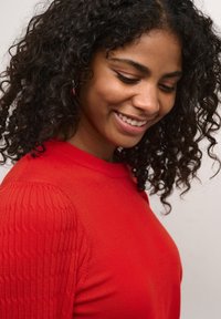 Pull en tricot rouge avec des manches côtelées texturées, un col rond et une silhouette ajustée, porté par une personne aux cheveux bouclés.