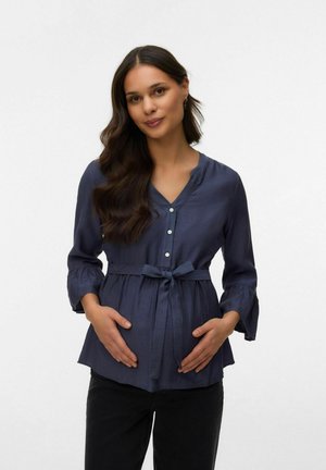 Ženska z dolgimi temnimi lasmi v mornarsko modri bluzi z zavezovanjem v pasu, ki drži roke na nosečniškem trebuhu, stoji pred enobarvnim ozadjem.