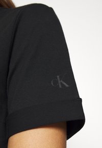 Black cotton t-shirt featuring a rolled sleeve and a subtle, contrasting "ck" logo printed on the sleeve. Soft texture, simple design.