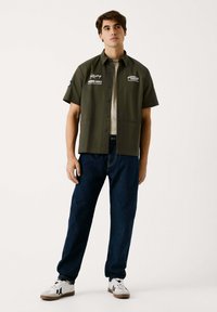 Camisa de manga corta verde oliva con cierre de botones, logotipos en el lado izquierdo del pecho, combinada con jeans de mezclilla azul oscuro y zapatillas blancas con detalles en negro.