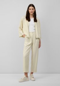 Lichtbeige, loszittende set van een jas en broek, met twee opgestikte zakken aan de voorkant. Gekleed met crèmewitte sneakers. Effen wit T-shirt eronder.