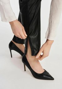 Czarne skórzane szpilki z ostrym noskiem, zestawione z czarnymi spodniami z rozcięciem przy kostce. Ręka dostosowująca nogawkę spodni.