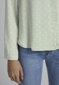 Blouse vert clair à pois blancs, manches longues et ourlet droit. Associée à un jean en denim bleu. Texture de tissu lisse.