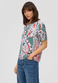 Krótki rękaw bluzki w zielonej bazie, ozdobionej kolorowym wzorem kwiatowym w odcieniach czerwieni, bieli i niebieskiego. Teksturowana ribbowa tkanina, luźny krój.