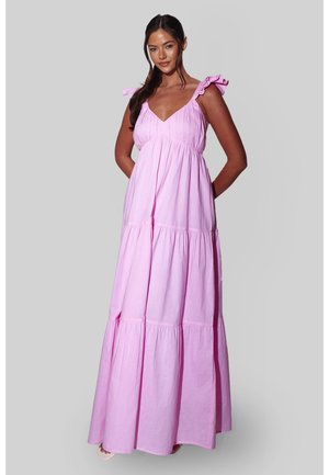 Kvinde med langt brunt hår iført en lys lyserød lagdelt maxikjole med flæseærmeløse stropper, stående mod en ensfarvet grå baggrund.