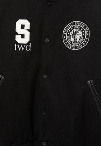 Sort uld baseballjakke med trykknapper, der har et hvidt "S" patch og broderet globuslogo. Inkluderer læderdetaljer på lommerne.