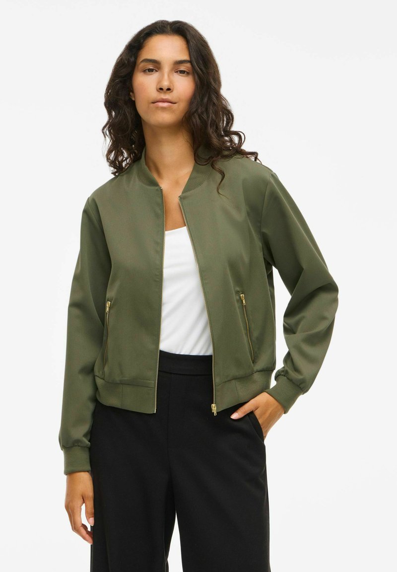 Olivengrüne Bomberjacke mit Reißverschluss vorne, gerippten Bündchen und zwei Seitentaschen. Hergestellt aus glattem, weichem Stoff. Kombiniert mit schwarzen Hosen.