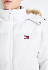 Giacca piumino bianca con collo alto, dotata di chiusura con zip e cappuccio con rifiniture in pelliccia. Patch con logo in rosso, bianco e blu sul petto.