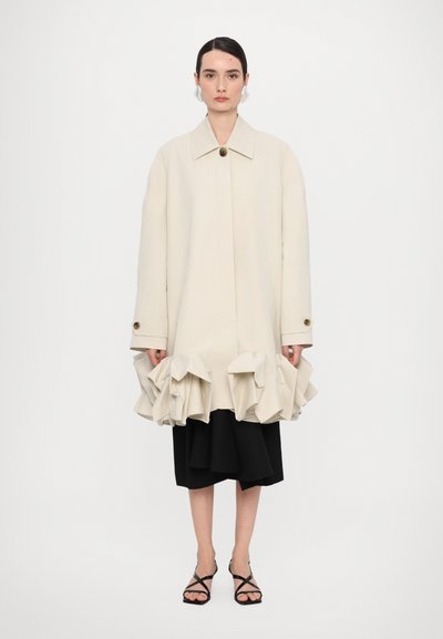 Manteau beige ajusté avec un ourlet large et volé, fermeture par un bouton unique, et coupe oversize, porté par-dessus une jupe noire à superpositions et des sandales à lanières.